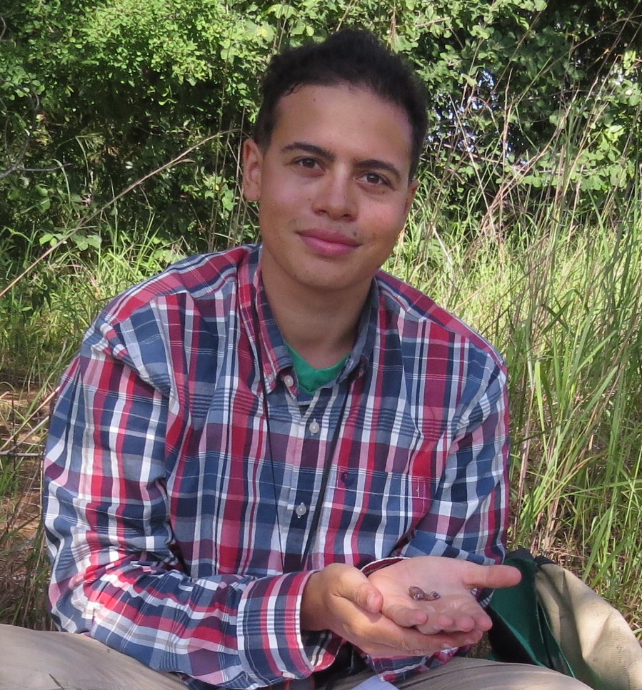 Jamie sitting in a field in a red and blue plaid shirt holding something in his palm and smiling at the camera