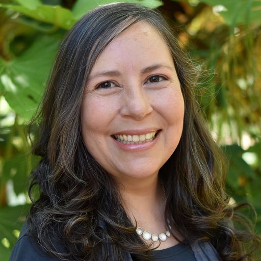 Dr. Briscoe smiling at the camera in front of a leafy green background. Her hair is brown and slightly curled and shoulder-length, and she wearsr a pearl necklace and dark blue top.