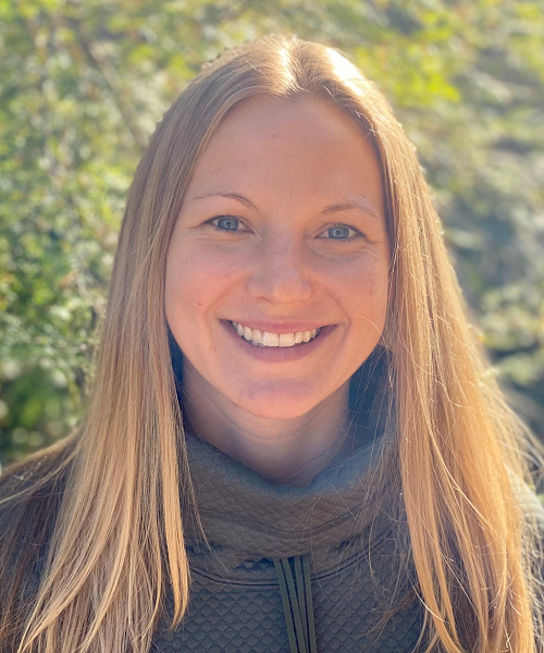 Megan DeMarche in a gray sweater in front of a leafy green background smiling at the camera