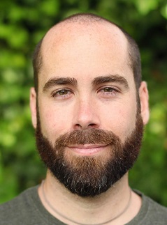 Moises with a beard and gray shirt smiling at the camera in front of a leafy green background.